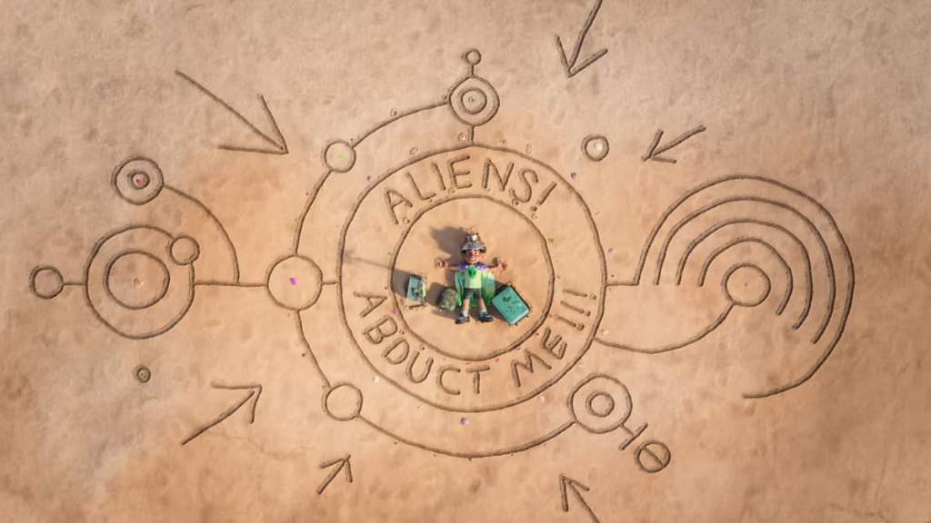 Elio laying in sand with the message "aliens abduct me" written around him