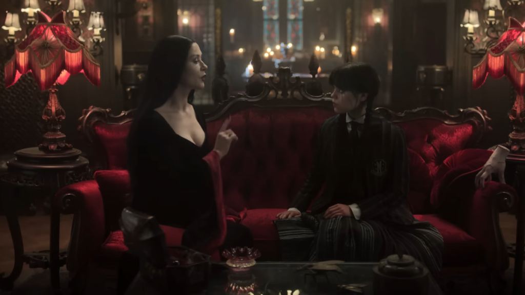 Morticia talking to Wednesday as they both sit on a dark red couch