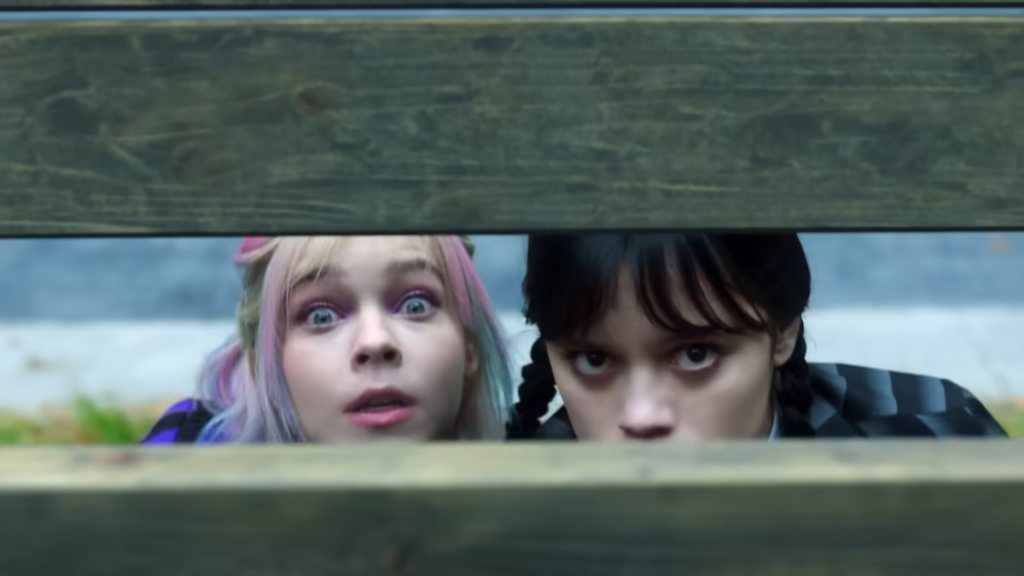 Enid and Wednesday peering through a wooden board