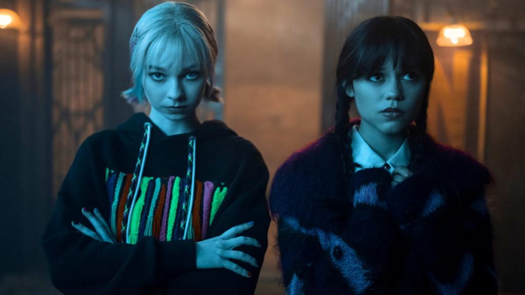 Enid standing with her arms crossed looking straight ahead with a serious expression next to Wednesday looking off to the side