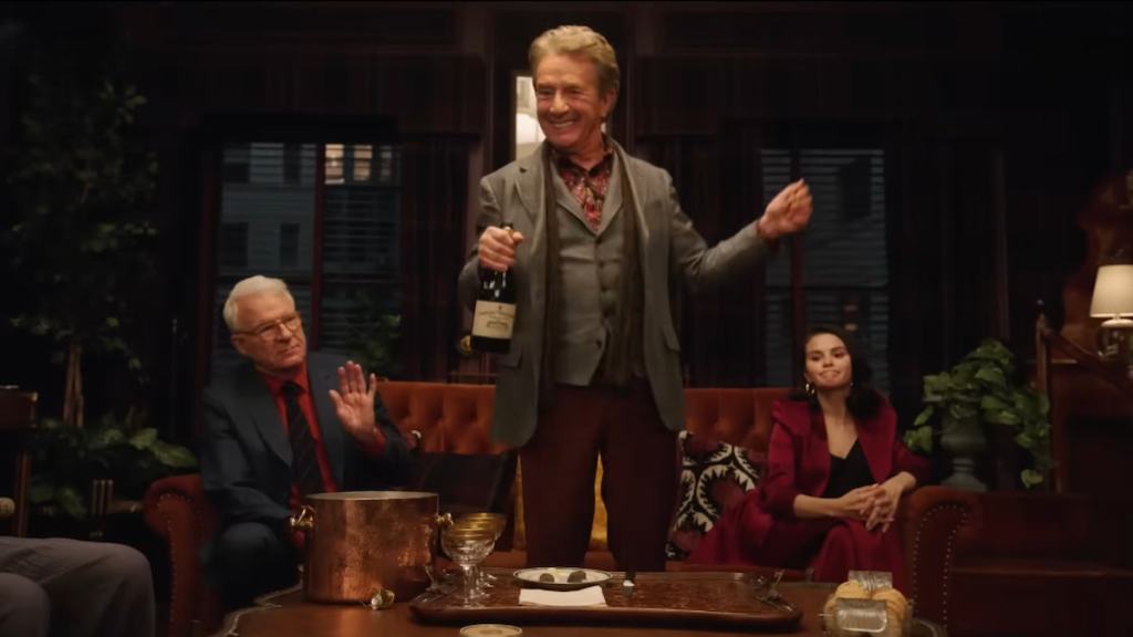 Oliver standing up with a drink and smiling while Charles and Mabel sit on a couch behind him in only murders in the building